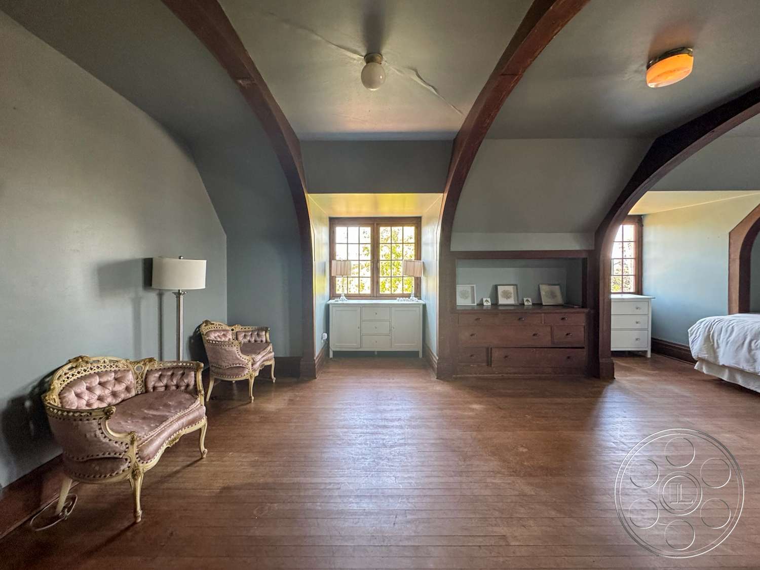 The Rosenheim Mansion - wooden dresser, vintage furniture, hardwood bedroom floors, decorative molding details, neutral painted walls, large windows with grid pattern, arched ceiling beams, pink upholstered armchairs, bright room lighting