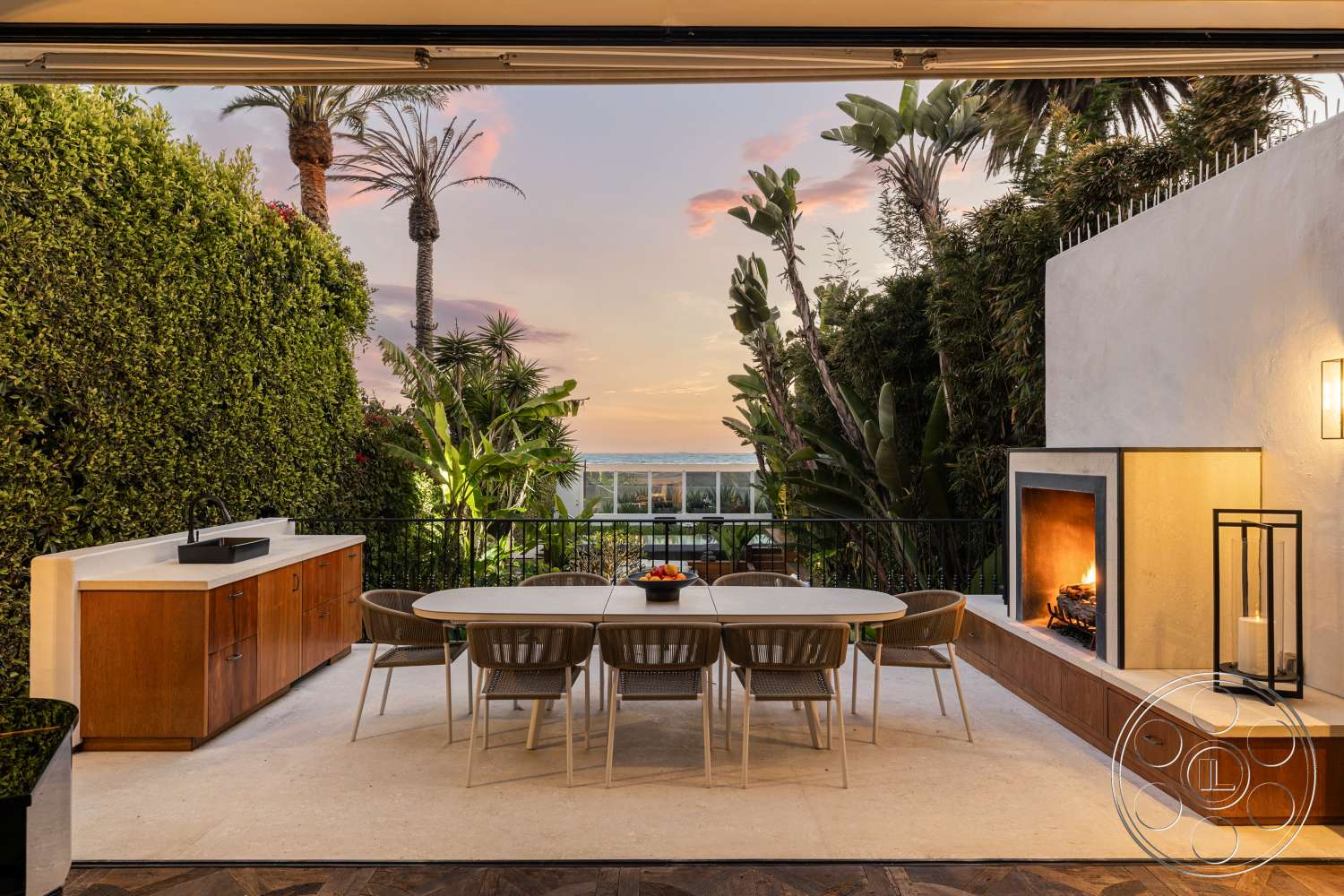 Beach 137 - exterior, palm trees, stone fireplace, landscaped greenery, al fresco dining area, modern architectural style, ocean view, ambient outdoor lighting, white stucco wall, woven dining chairs, concrete patio flooring, wooden outdoor dining table, built-in outdoor kitchen cabinetry, level patio space