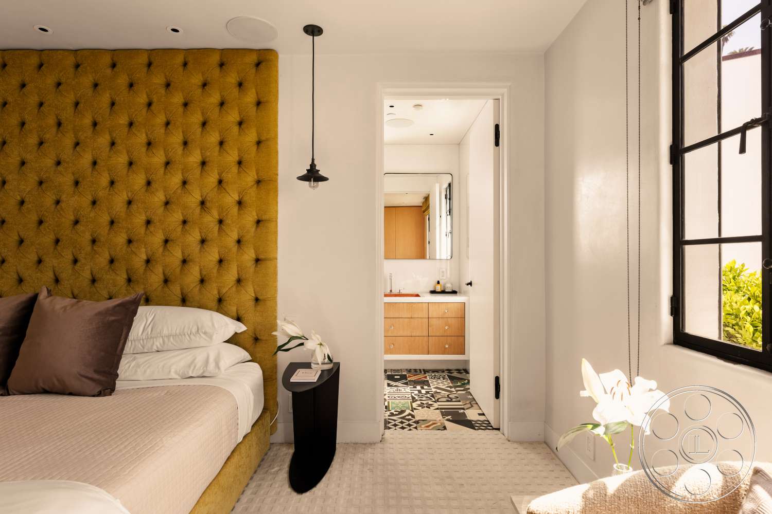 Beach 137 - dark hardwood floor, black framed window, patterned tile flooring, metal pendant lighting, mirrored bathroom wall, wood vanity with sink, textured white walls, decorative flower arrangement, tufted yellow velvet headboard, painted white door, neutral bedding with brown throw pillows, small black side table