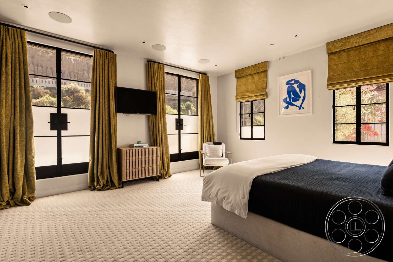 Beach 137 - abstract wall art, TV mounted on wall, wood bedside table, textured carpet flooring, white painted walls, light neutral color palette, large windows with black frames, contemporary bedroom, pleated fabric curtains, low-profile bed with dark bedding, soft accent chair, decorative window treatments