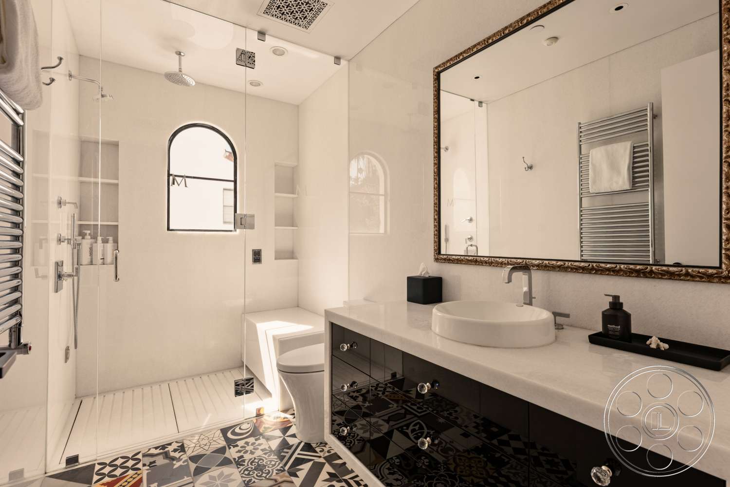 Beach 137 - open shelving, black cabinetry, marble countertop, arched window, glass shower doors, ornate framed mirror, spotlighting ceiling lights, modern plumbing fixtures, white wall tiles, white floating vanity, rectangular vessel sink, colorful patterned floor tiles, chrome towel warmer, toilet with hidden tank