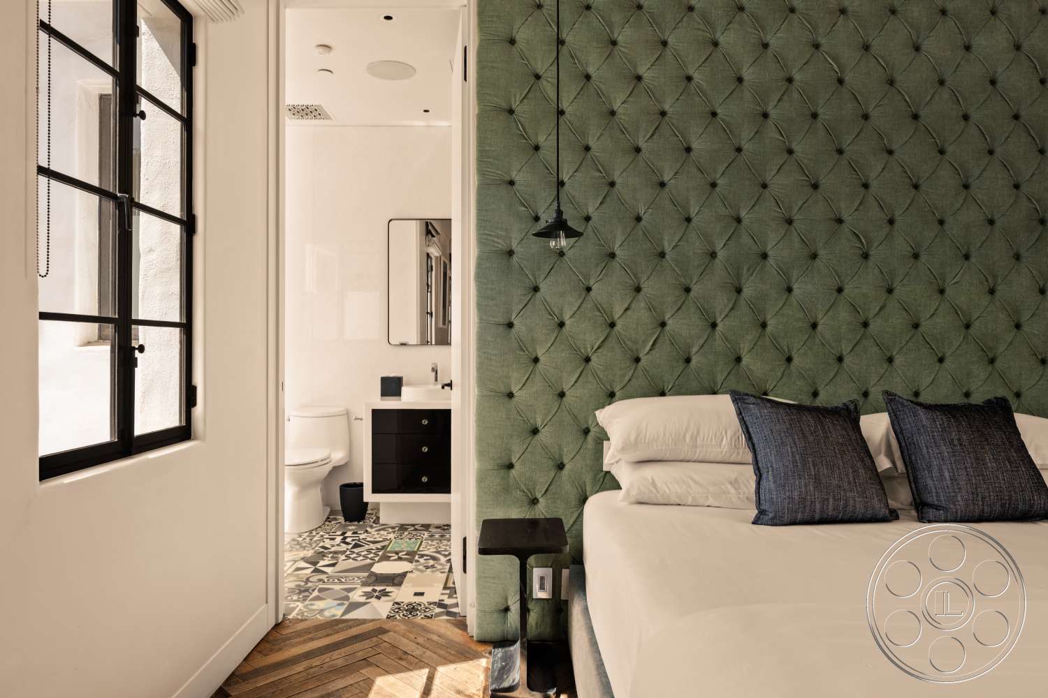 Beach 137 - minimalist design elements, black metal window frames, white painted walls, floor-to-ceiling window view, tufted green fabric headboard, hardwood herringbone bedroom flooring, geometric patterned tile bathroom flooring, modern bathroom vanity with black cabinetry, pendant light fixture above nightstand, dark blue decorative pillows