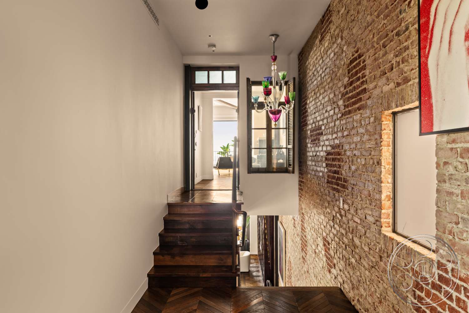 Beach 137 - open floor plan, natural light from large windows, modern eclectic design, interior hallway, white painted walls, exposed red brick accent wall, hardwood stairs, herringbone pattern hardwood flooring, multicolored glass chandelier, arched entryway with glass doors