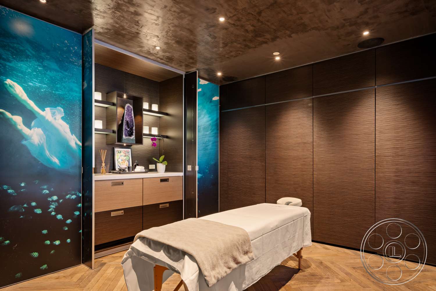 Beach 137 - dark wood cabinetry, floating shelves, soft ambient lighting, concrete ceiling, wood herringbone flooring, textured wooden wall panels, modern massage room, spa decor elements, water-themed wall art
