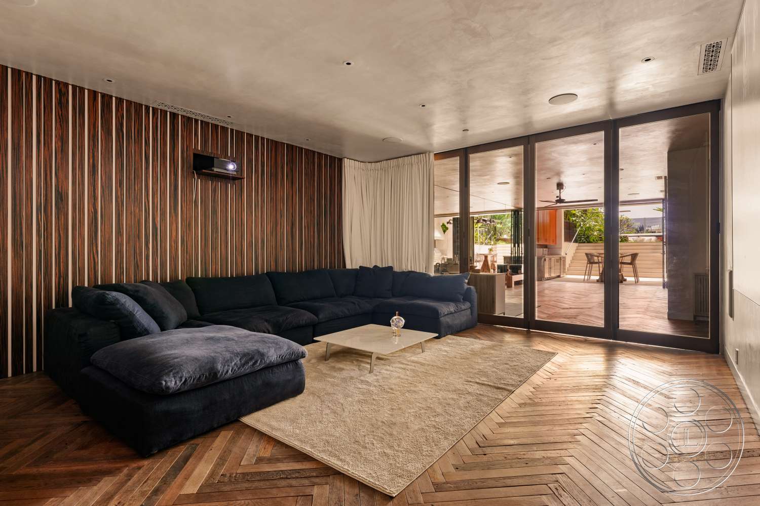 Beach 137 - open floor plan, indoor-outdoor flow, floor-to-ceiling windows, neutral area rug, dark wood accent wall, large sliding glass doors, minimalist design aesthetic, chevron hardwood flooring, plaster ceiling, sectional sofa with plush cushions, integrated projector