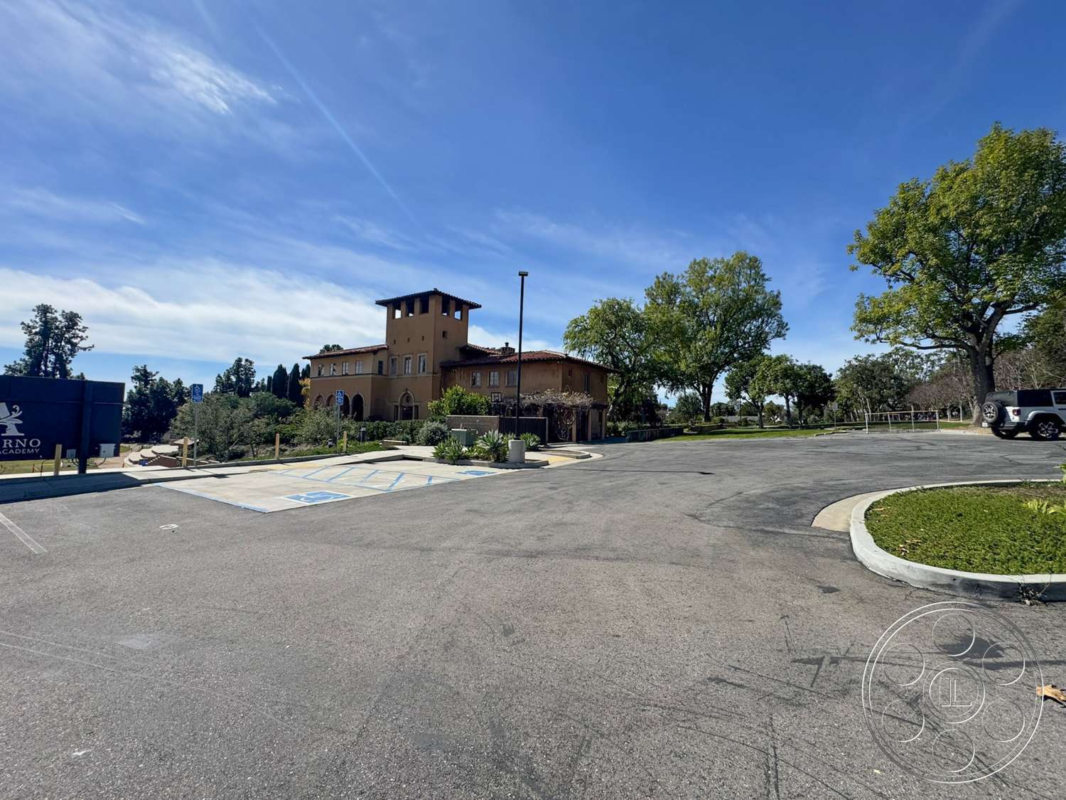 School 20 - exterior, landscaped garden, large trees, open outdoor space, curved driveway, terracotta roof tiles, arched doorways, signage, Mediterranean architectural style, two-story building, concrete parking lot, stucco exterior walls, blue sky with scattered clouds, institutional setting