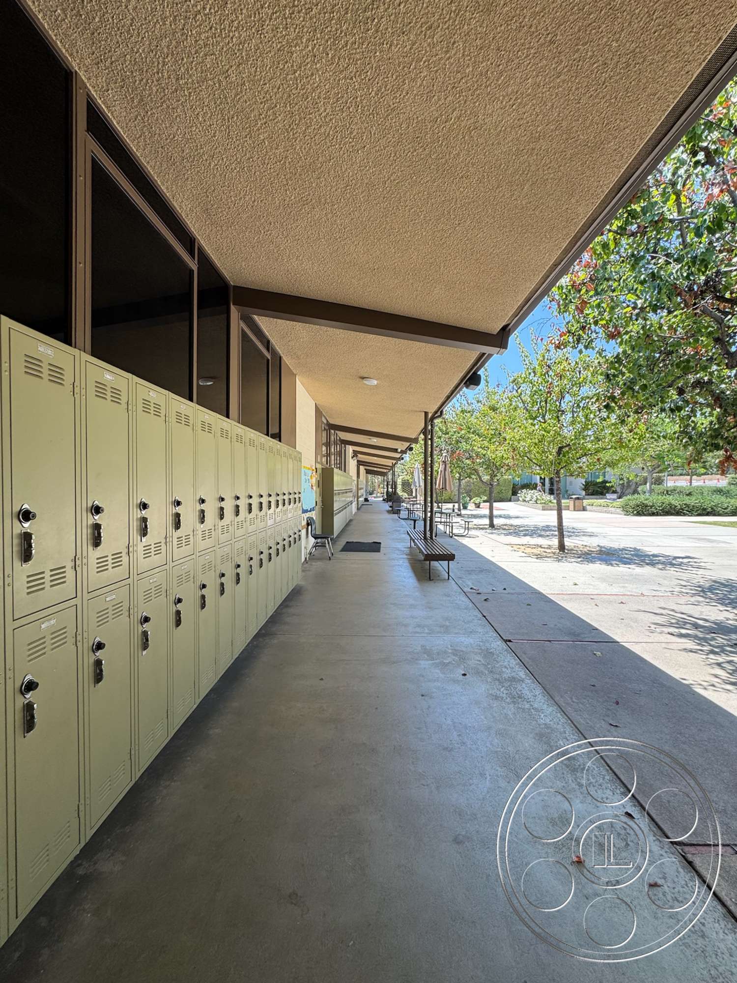 School 20 - exterior, concrete walkway, metal lockers, flat roof overhang, landscaped outdoor area, exposed aggregate concrete surface, trees lining the walkway, green lockers, clearly visible seating area, modern school architecture