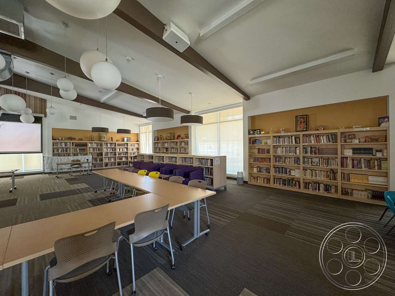 School 20 - open layout, modern aesthetic, vaulted ceiling with exposed wood beams, rectangular conference tables, colorful accent chairs, soft white pendant lighting, contemporary style furniture, wooden bookshelves with books, carpet flooring with textured pattern, large windows with sheer coverings, warm-toned accent wall, library reading area
