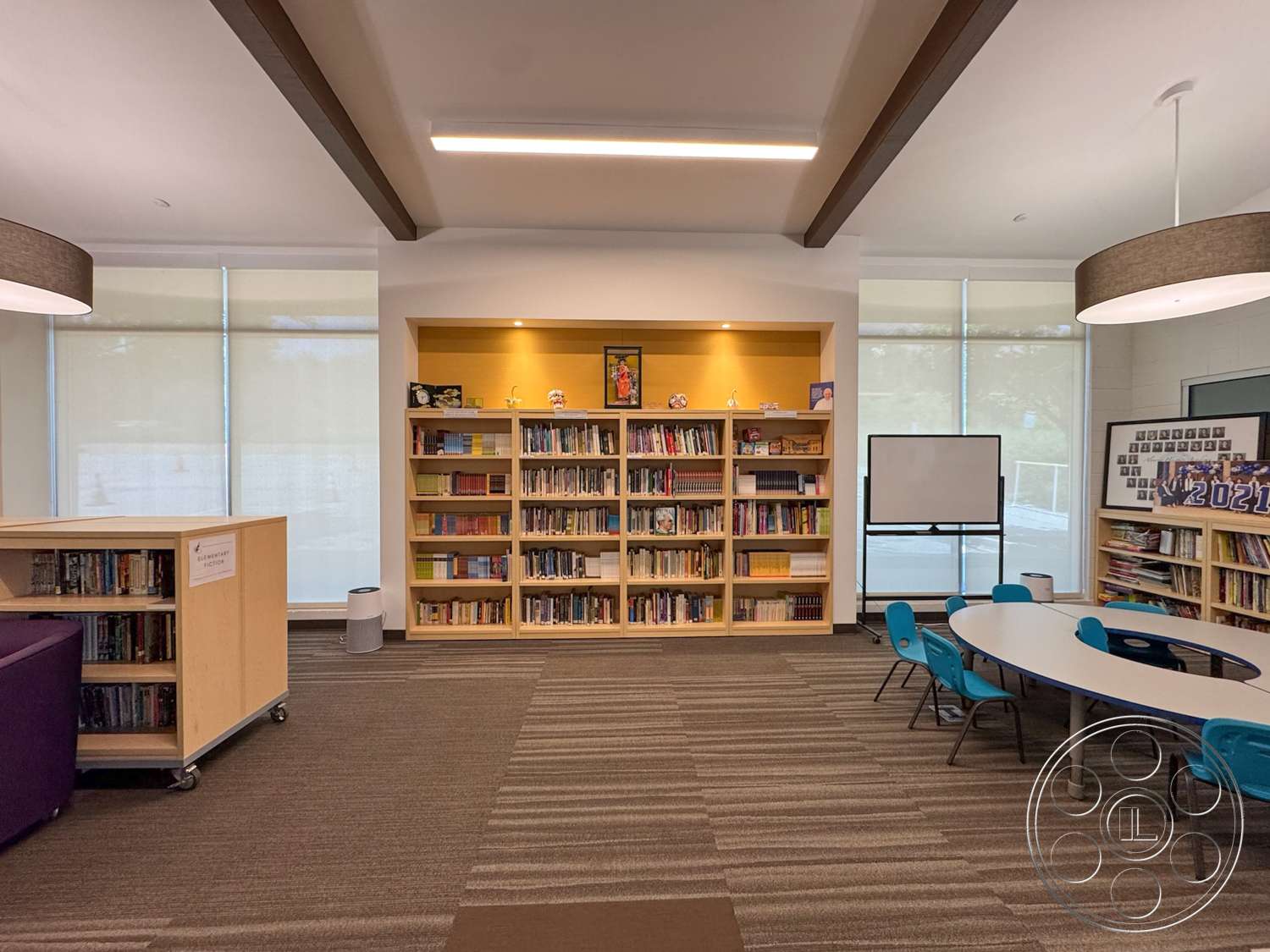 School 20 - white walls, open layout, carpet flooring, exposed wood ceiling beams, textured carpet tiles, linear LED ceiling lights, built-in wooden bookshelves, blue plastic chairs, yellow painted wall accent, round collaborative study table, large windows with sheer shades, multifunctional classroom space