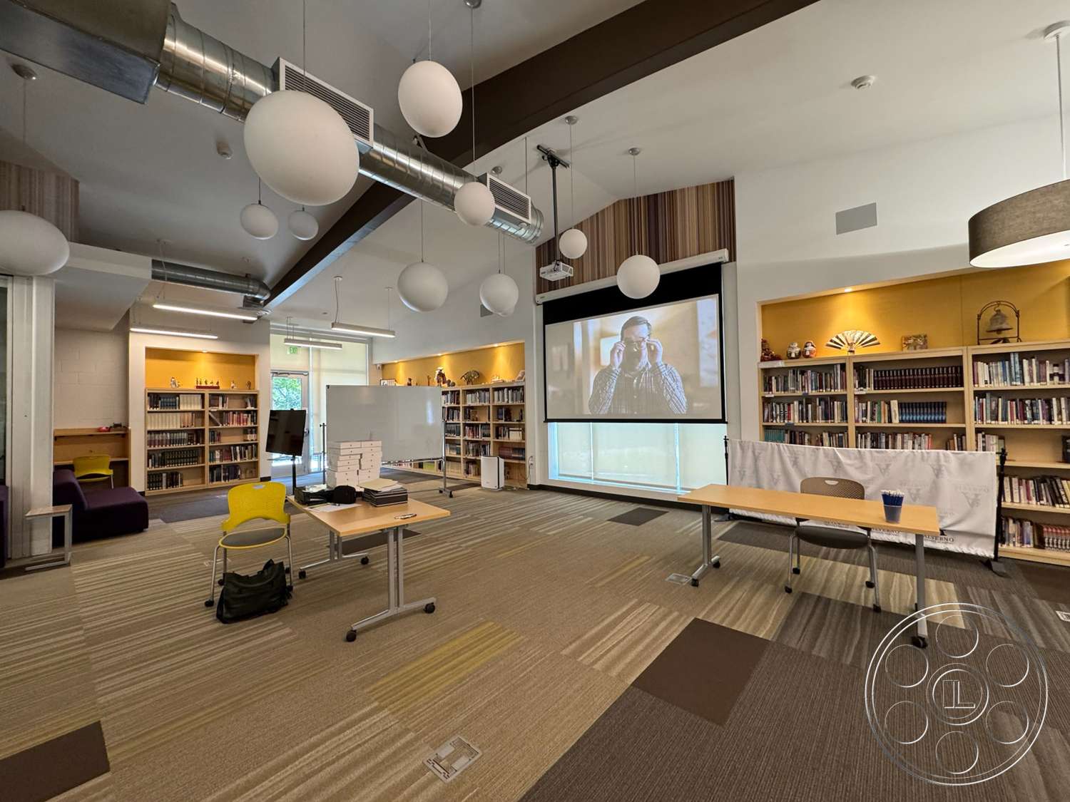 School 20 - contemporary furniture, open floor plan, modern aesthetic, glass windows, neutral wall color, exposed ductwork, textured carpet flooring, wood beam ceiling, linear suspended ceiling lights, wall-mounted projection screen, built-in library shelving, large whiteboard area, multicolored lounge seating, yellow chair seating