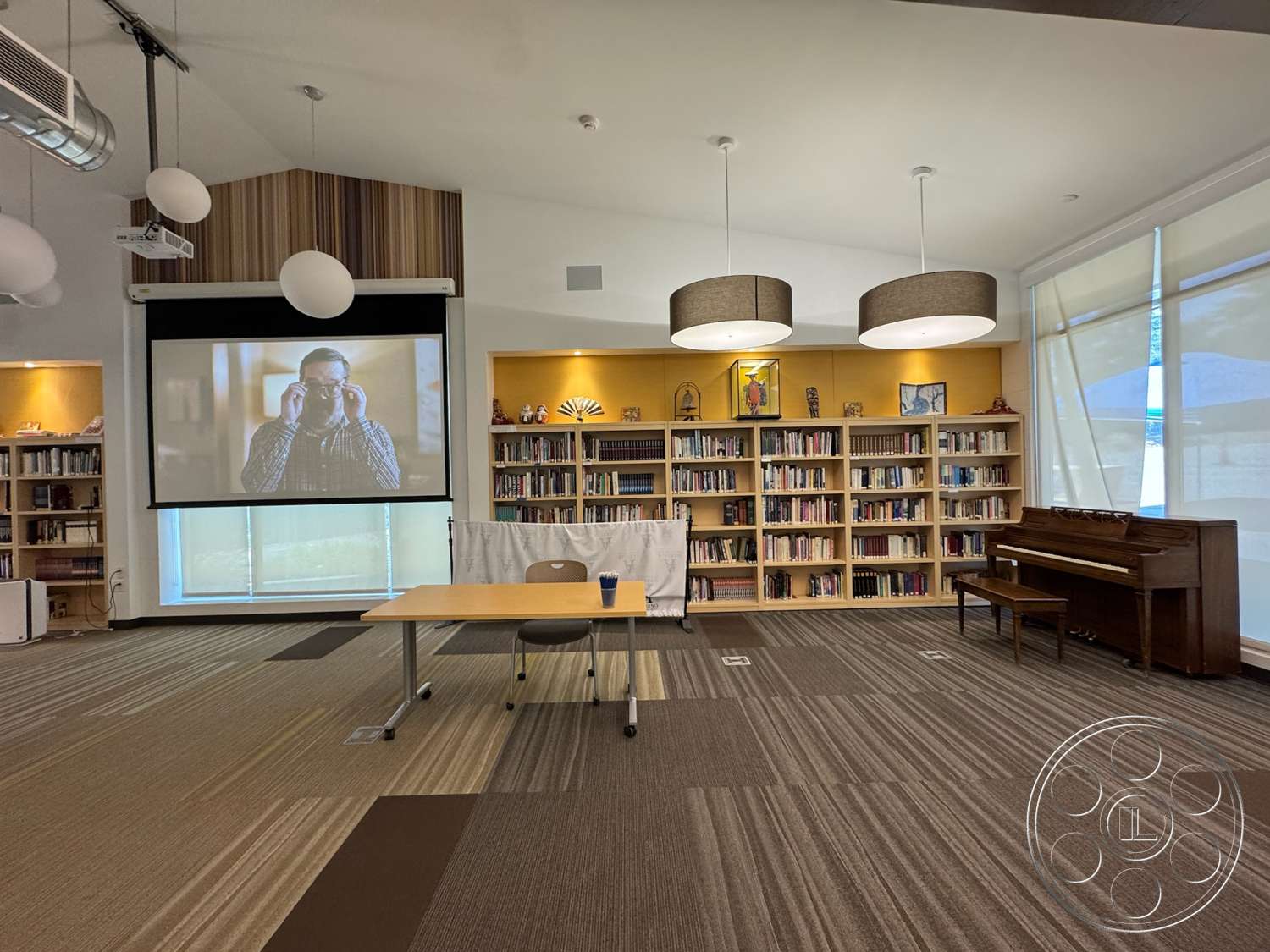 School 20 - carpet flooring with striped pattern, wooden bookshelf with open shelves, drop ceiling with circular light fixtures, projector screen mounted on wall, piano in wooden finish, long rectangular table with chairs, large windows with sheer blinds, acoustic panels on wall, yellow accent wall with decorative items