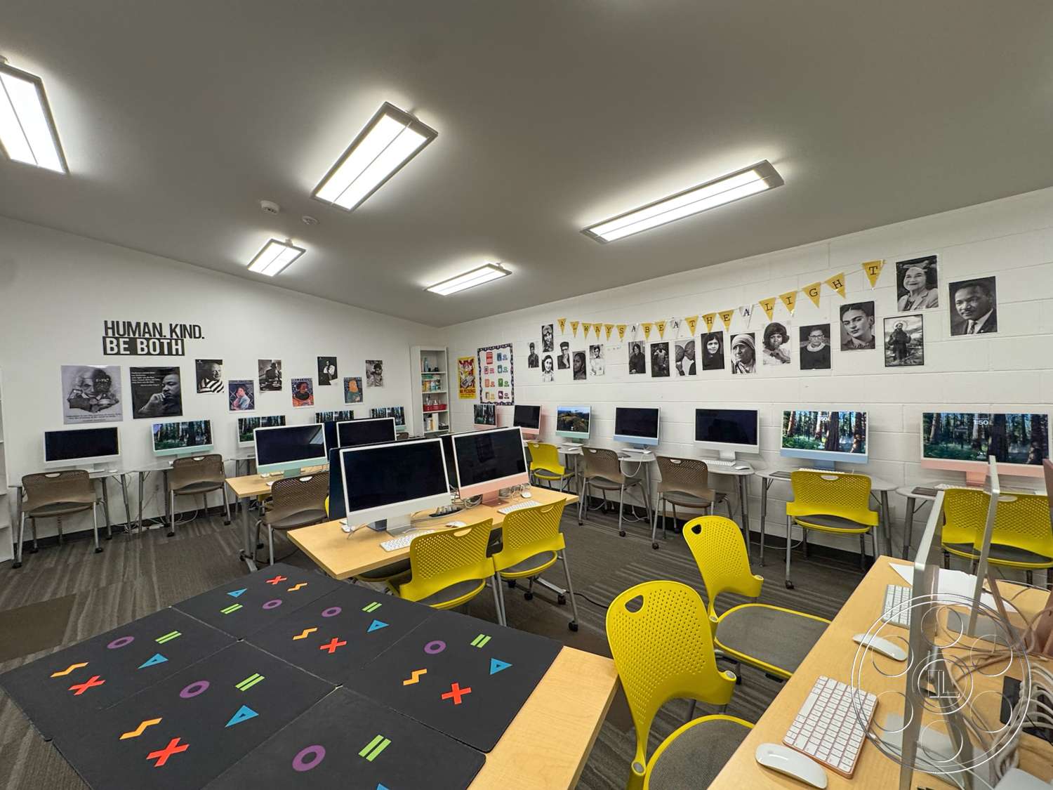 School 20 - white painted walls, drop ceiling with fluorescent lighting, shelves for storage, large wall-mounted computer workstations, light brown carpet flooring, yellow plastic chairs with perforated design, wooden tables for computer work, graphic wall art and posters, decorative banner with triangular flags, open layout classroom