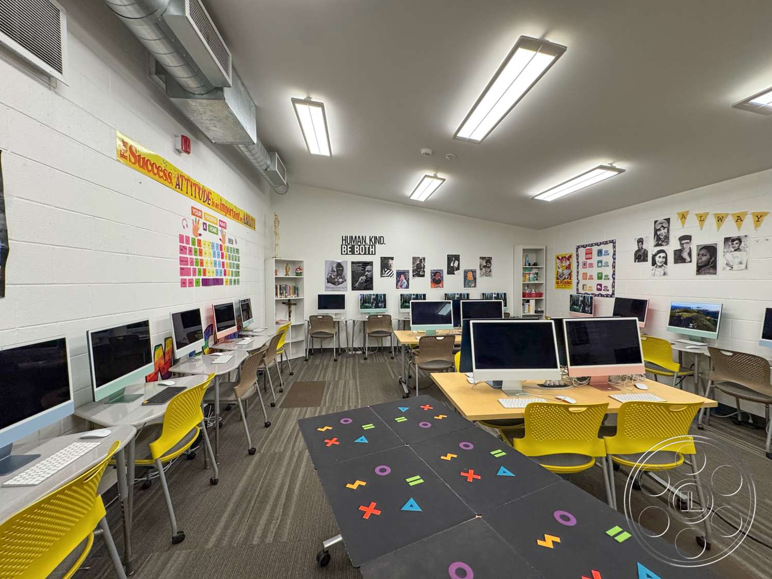 School 20 - carpet flooring, exposed ductwork ceiling, fluorescent ceiling lights, light gray painted walls, yellow plastic chairs, computer desks with silver legs, black table with colorful designs, wall-mounted display boards, educational posters, computers with flat-screen displays