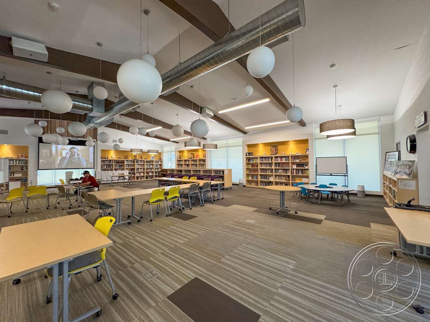 School 20 - floor-to-ceiling windows, metal ductwork, pendant lighting fixtures, exposed wood ceiling beams, wooden bookshelves, carpeted flooring with striped pattern, modular study tables, ergonomic yellow chairs, media screen, interior learning space, open concept reading area