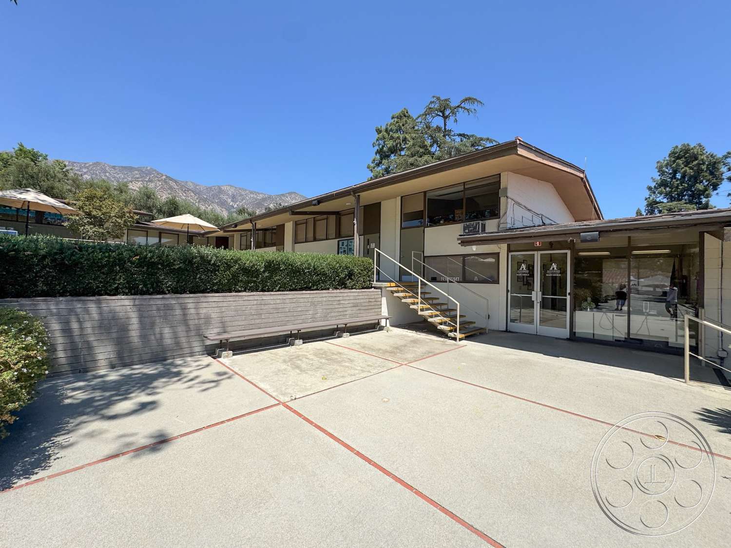 School 20 - exterior, modern architectural style, concrete walkway, mountain backdrop, hedge landscaping, concrete patio floor, glass double doors, open courtyard setting, flat roof with overhang, wooden stairs with metal railing
