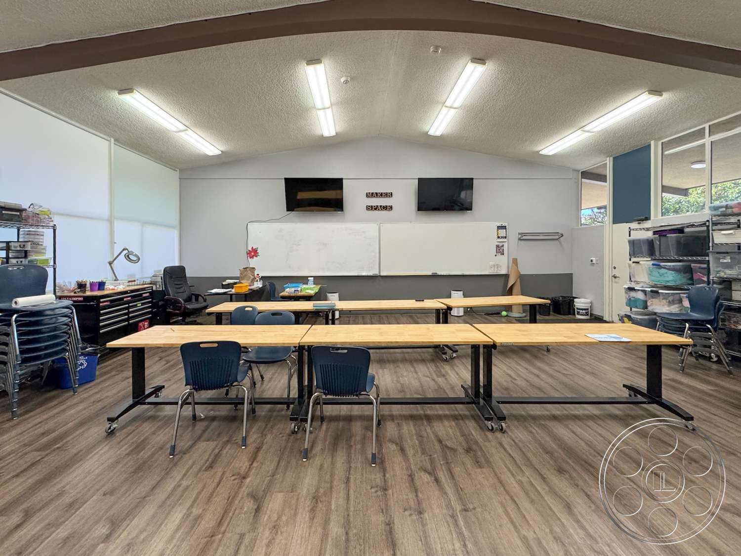 School 20 - storage cabinets, laminate wood flooring, vaulted ceiling with exposed beams, large windows with natural light, whiteboard wall, rectangular classroom layout, metal-framed tables, plastic stacking chairs, LED fluorescent lighting, tv wall mount, art supplies area