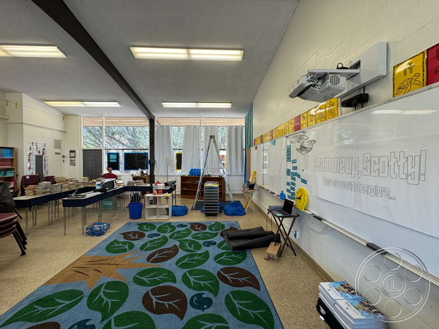 School 20 - tile flooring, open layout, storage cabinets, textured plaster walls, metal folding chairs, drop ceiling with fluorescent lights, large windows with sheer curtains, interior classroom, wooden teacher's desk, colorful educational wall displays, multi-colored leaf-patterned area rug, projector mounted on wall, whiteboard with educational graphics
