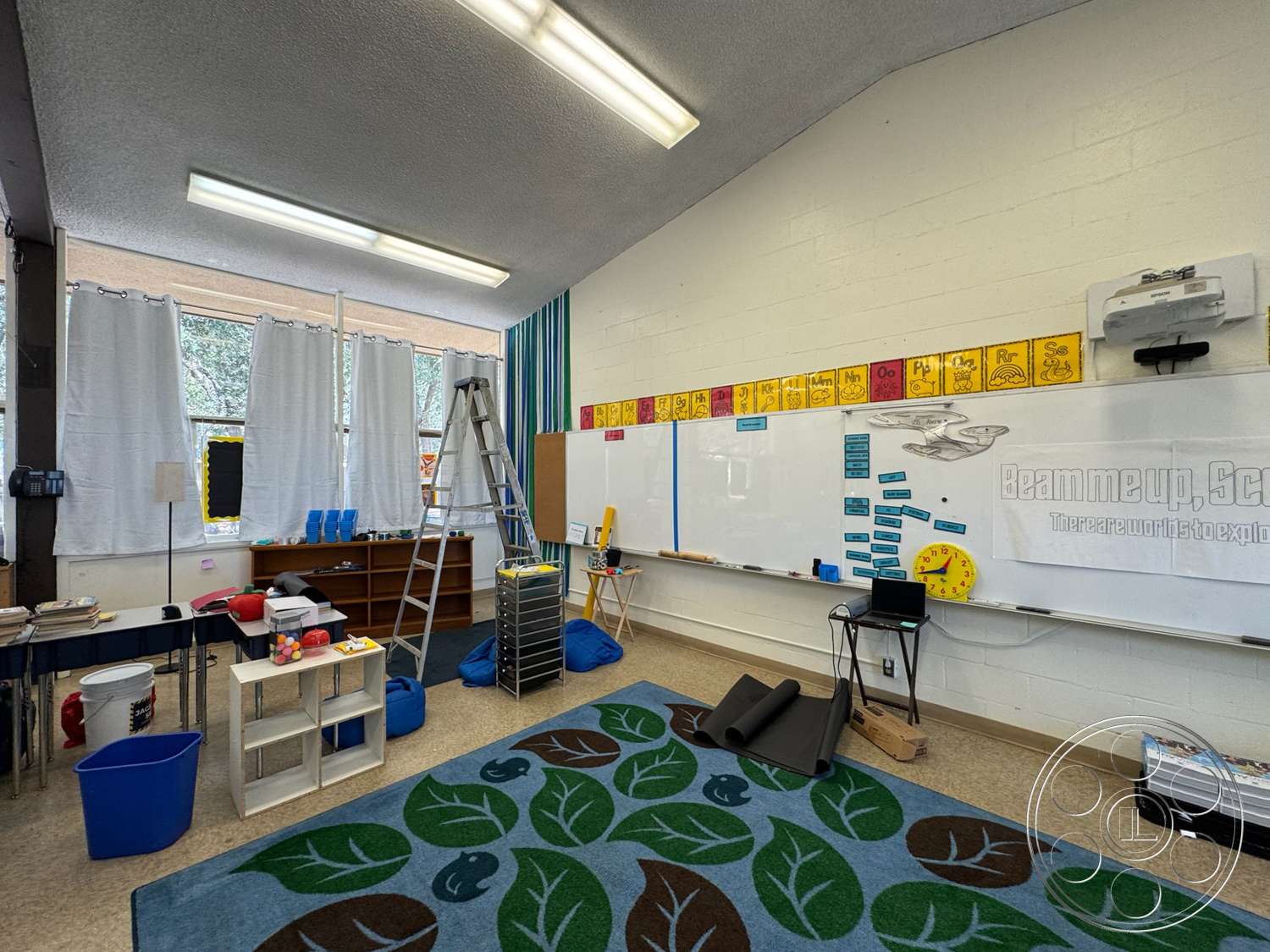 School 20 - wooden bookshelf, colorful storage bins, acoustic ceiling tiles, painted cinder block walls, interior classroom space, dry erase board, laminate classroom flooring, window treatments with sheer curtains, multicolored educational wall decorations, ladder for accessibility, folding table for classroom activities, cube storage unit, blue bean bag seating, decorative area rug with leaf pattern, hanging projector, task lighting with fluorescent fixtures