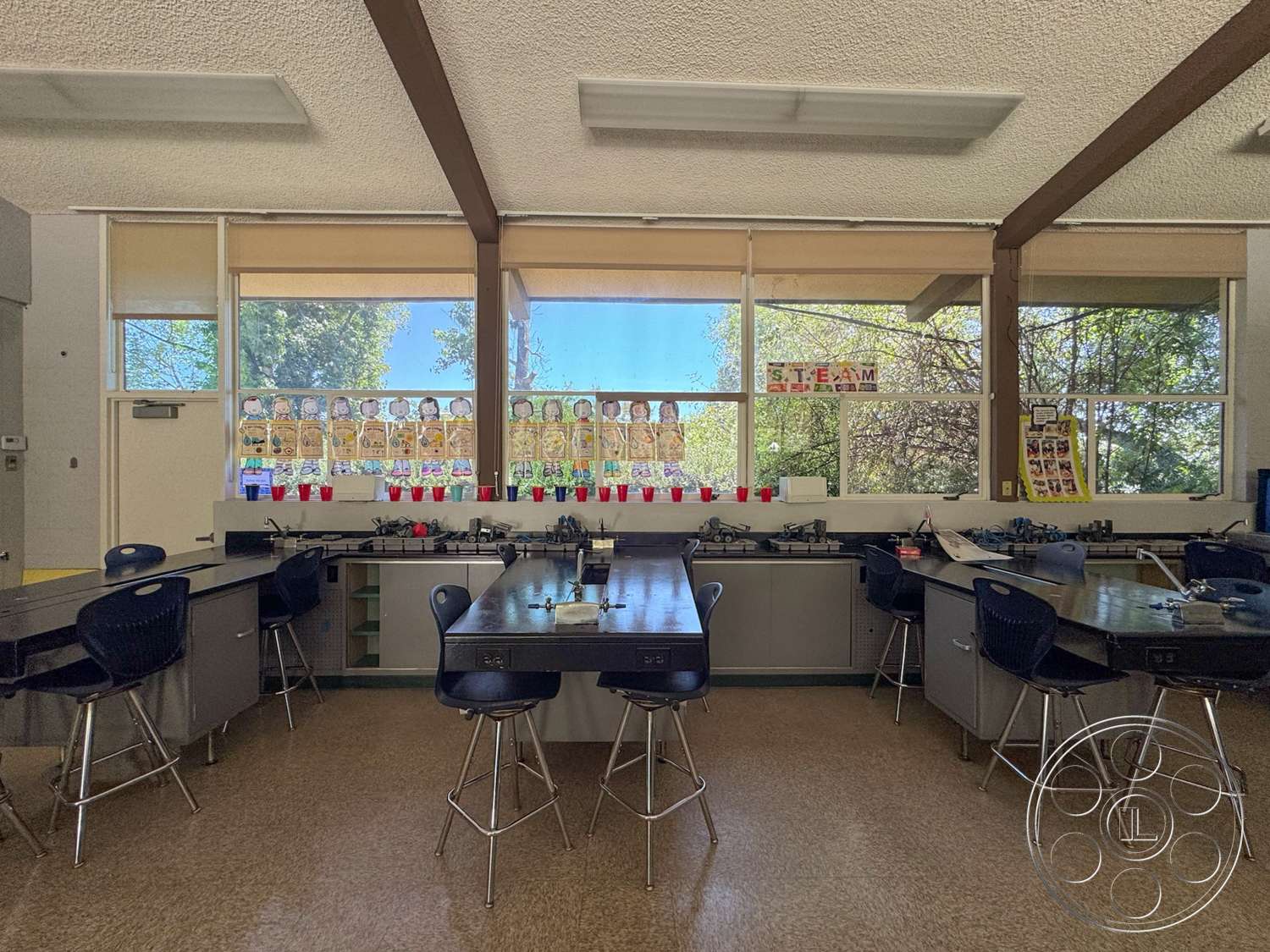 School 20 - laminate countertops, wood beam ceiling, textured ceiling panels, large windows with blinds, open classroom layout, vinyl floor tiles, metal laboratory tables, educational wall displays, colorful plastic cups, students' artwork display