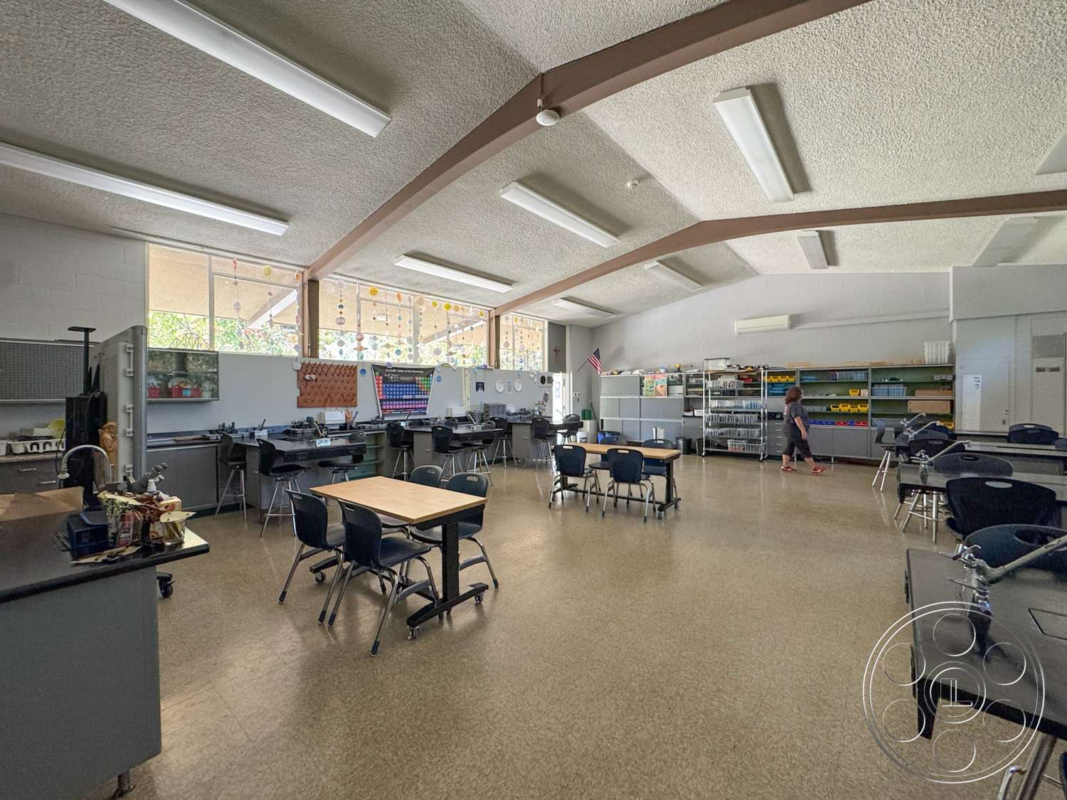 School 20 - metal shelving units, glass windows, vinyl flooring, grey painted walls, black plastic chairs, acoustic ceiling tiles, exposed wood ceiling beams, large windows with natural light, fluorescent ceiling lights, interior classroom, rectangular classroom tables, art supply storage, colorful decorative items, dual-purpose room