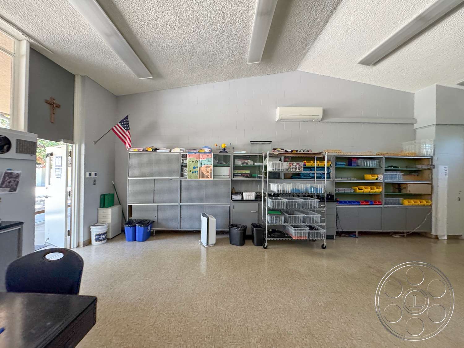School 20 - large windows for natural light, metal shelving unit, textured plaster walls, American flag, vaulted ceiling with exposed beams, plastic storage bins, white-painted walls, linoleum flooring, signage on wall