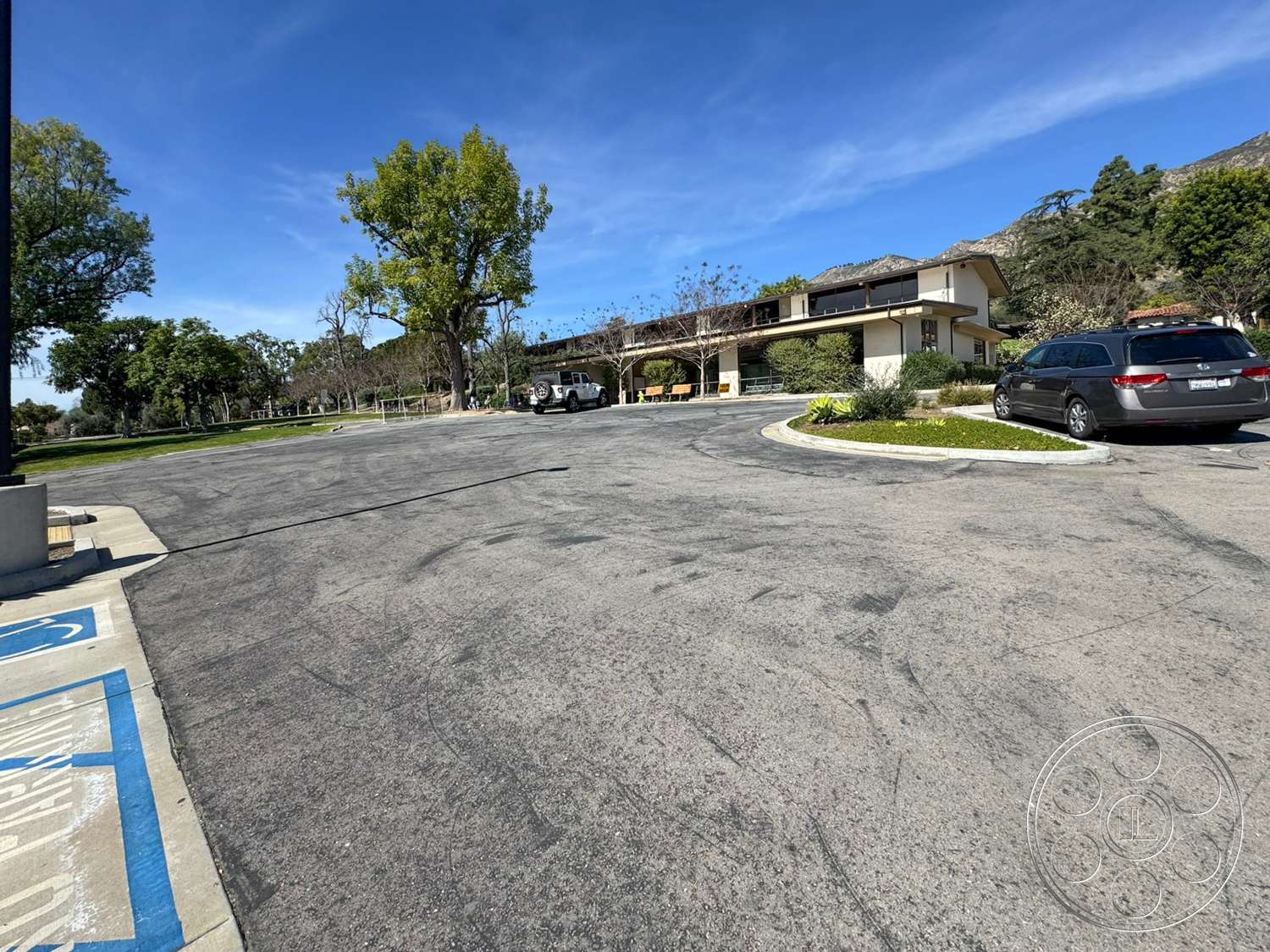 School 20 - metal railing, glass windows, curved driveway, mountain backdrop, blue sky, asphalt parking lot, leafy trees, landscaped garden area, exterior stone planters, two-story contemporary building