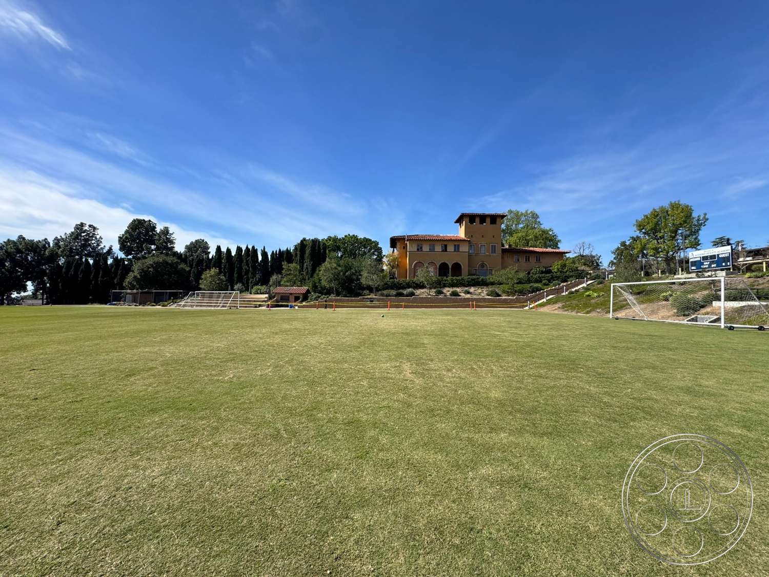 School 20 - exterior, landscaped garden, outdoor seating area, surrounding greenery, paved walkways, manicured lawn, Mediterranean architecture, red tile roof, arched doorways, soccer field, tan stucco walls, cedar trees, multi-story home