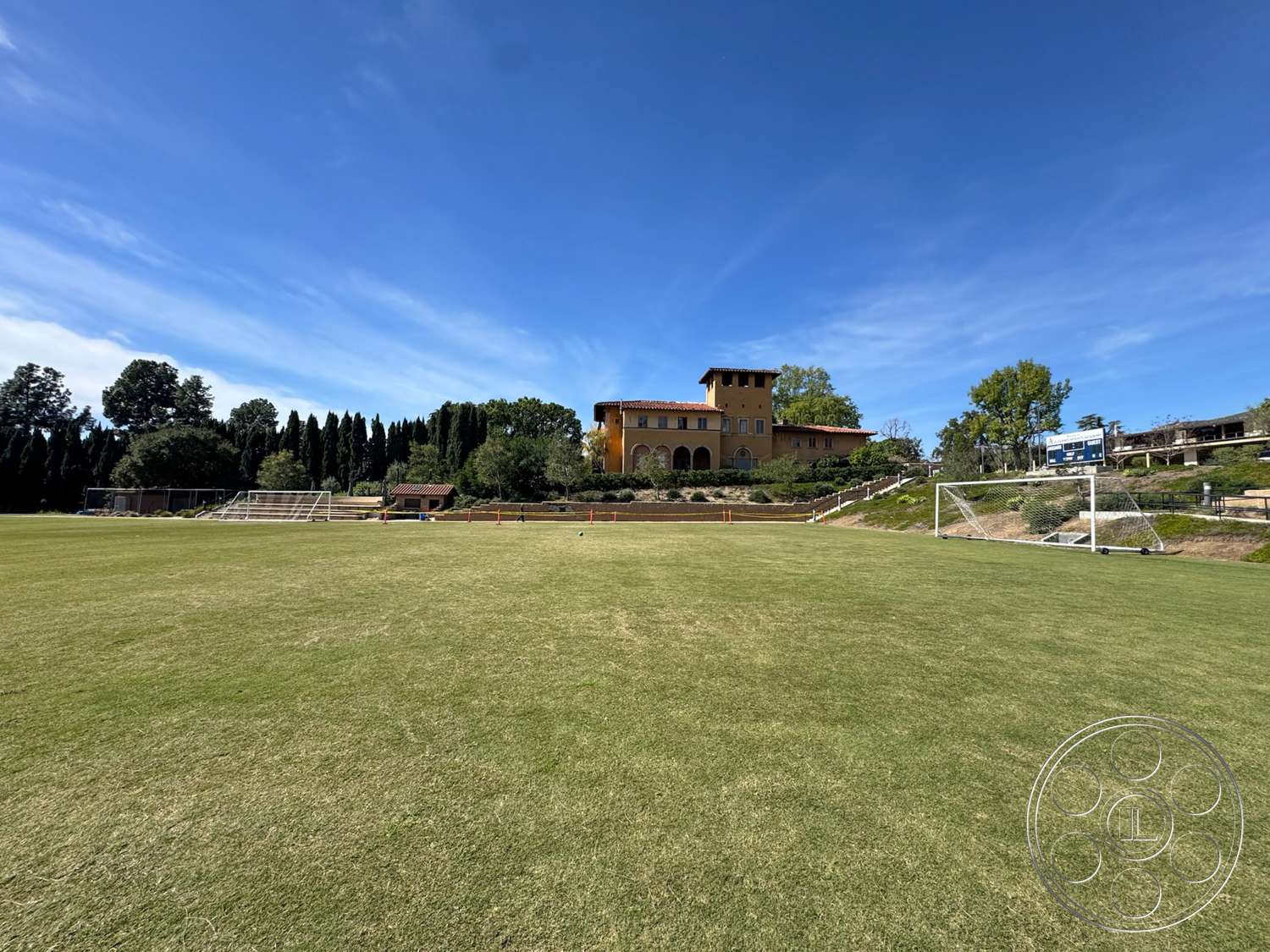 School 20 - exterior, landscaped gardens, surrounding trees, concrete walkway, red tile roof, stucco walls, arched windows, Mediterranean architectural style, goalposts, green grass field, balcony with iron railing