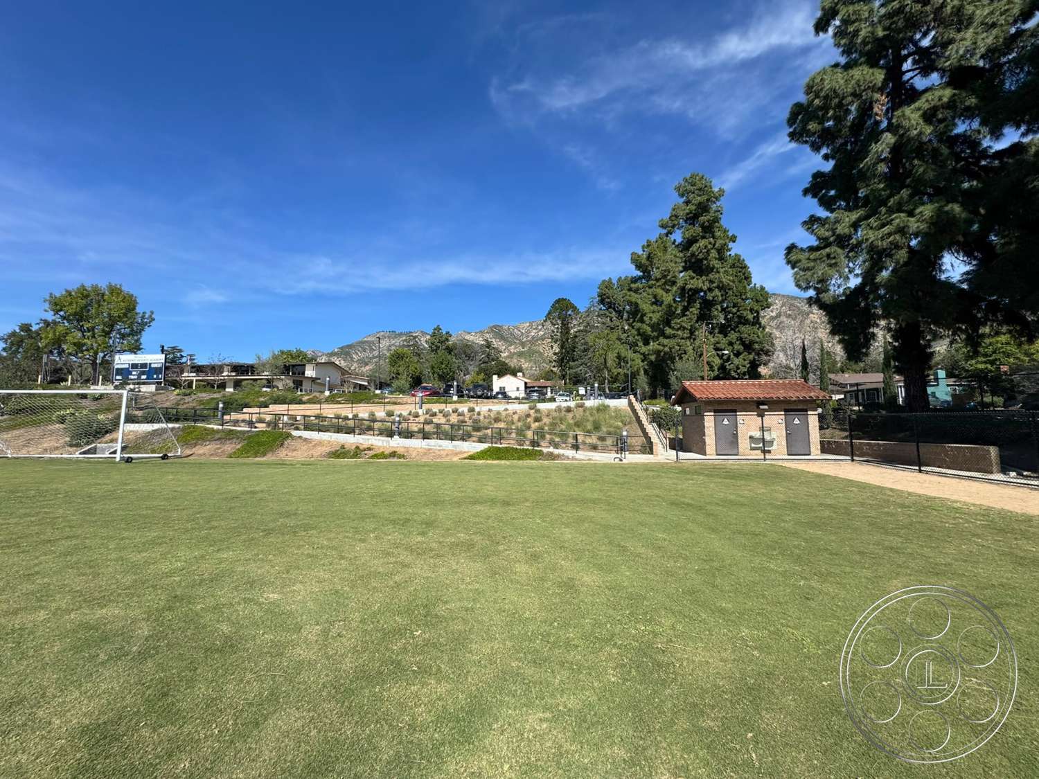 School 20 - exterior, mountain backdrop, clear blue sky, tall evergreen trees, park setting, natural grass soccer field, landscaped hillside with terraced planting beds, brick restroom building with red tile roof, chain-link fence surrounding the field
