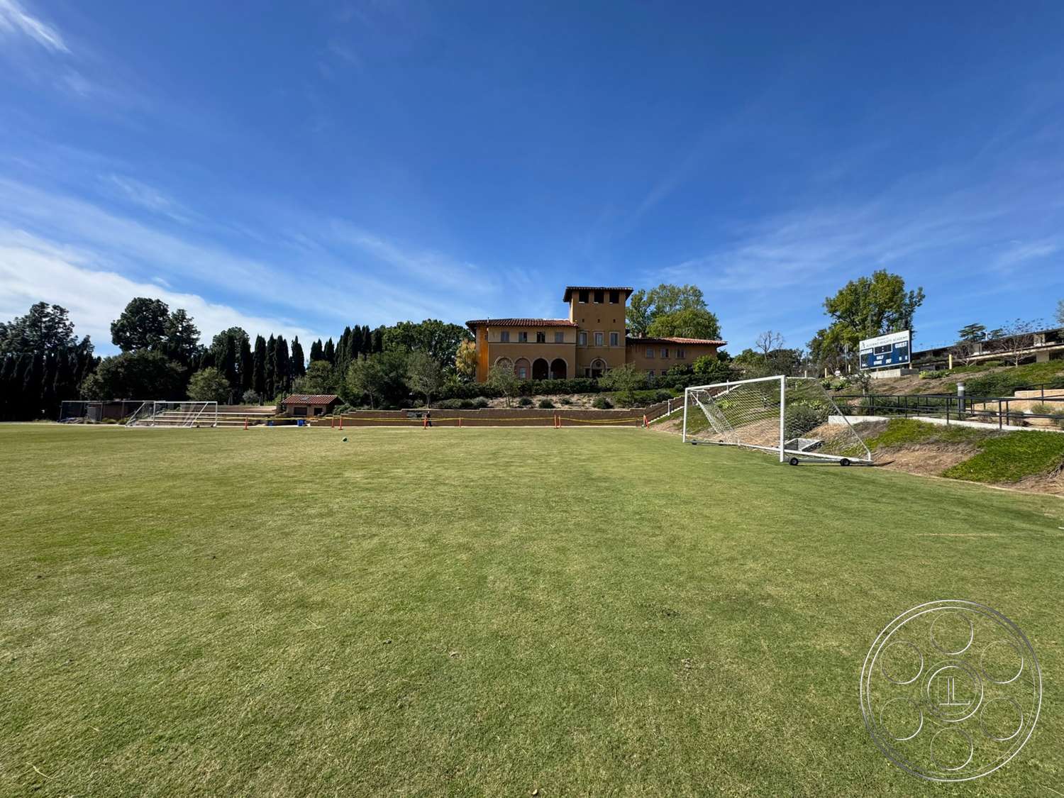 School 20 - large windows, exterior, open outdoor space, manicured lawn, clear blue sky, red tile roof, arched doorways, stone retaining wall, Mediterranean architectural style, yellow stucco walls, multi-story home, landscape with trees and shrubs, soccer field with goals