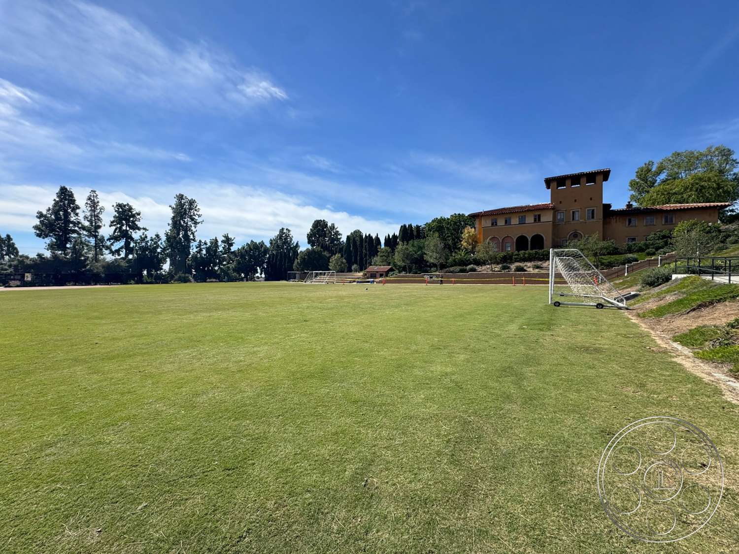 School 20 - exterior, landscaped gardens, stucco walls, tile roof, arched windows, tree line, Mediterranean architectural style, goalposts, sunny outdoor setting, green grass field, built stone structure