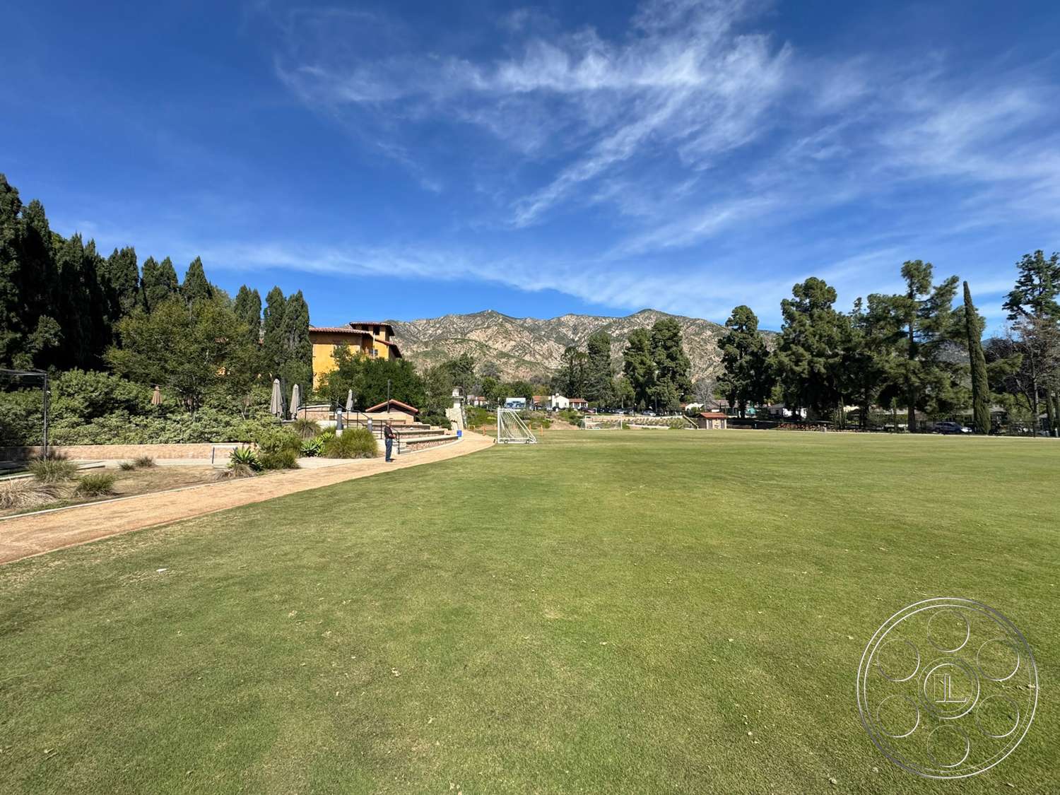 School 20 - exterior, surrounding greenery, mountain backdrop, landscaped path, terraced garden beds, urban park setting, grassy lawn area, sunny clear sky, tree line border, hardscape seating area, yellow stucco building