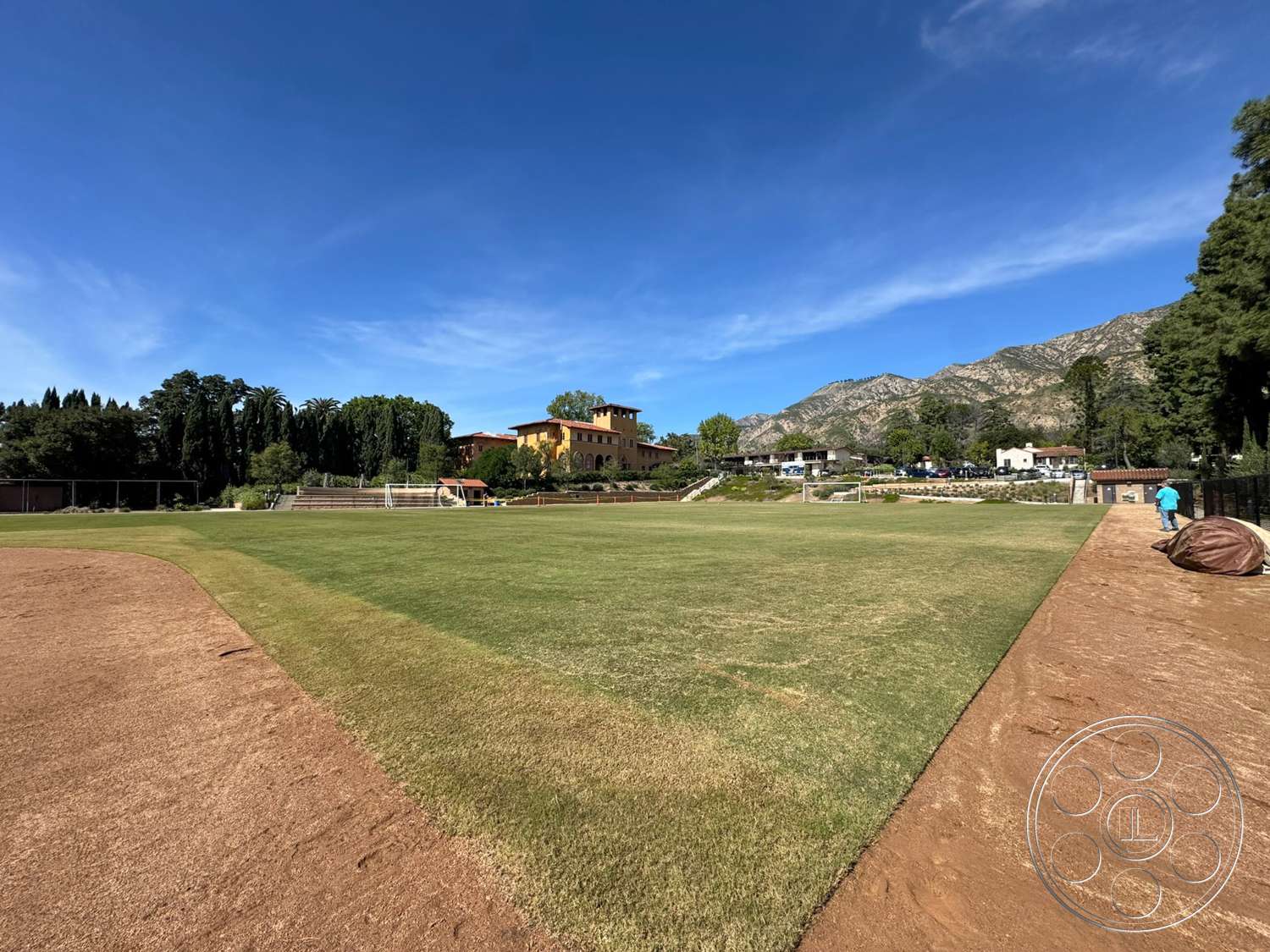 School 20 - exterior, landscaped gardens, outdoor seating area, paved pathways, stucco facade, two-story building, concrete retaining wall, surrounding mountain view, grass athletic field, dirt infield, red-tiled roof, row of trees