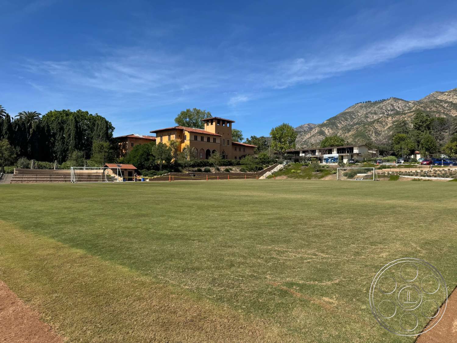 School 20 - exterior, landscaped garden, concrete pathway, mountain backdrop, terracotta roof tiles, tall palm trees, open sky, stucco exterior walls, wooden seating area, grass sports field, two-story Mediterranean-style building, photosynthetic green lawns