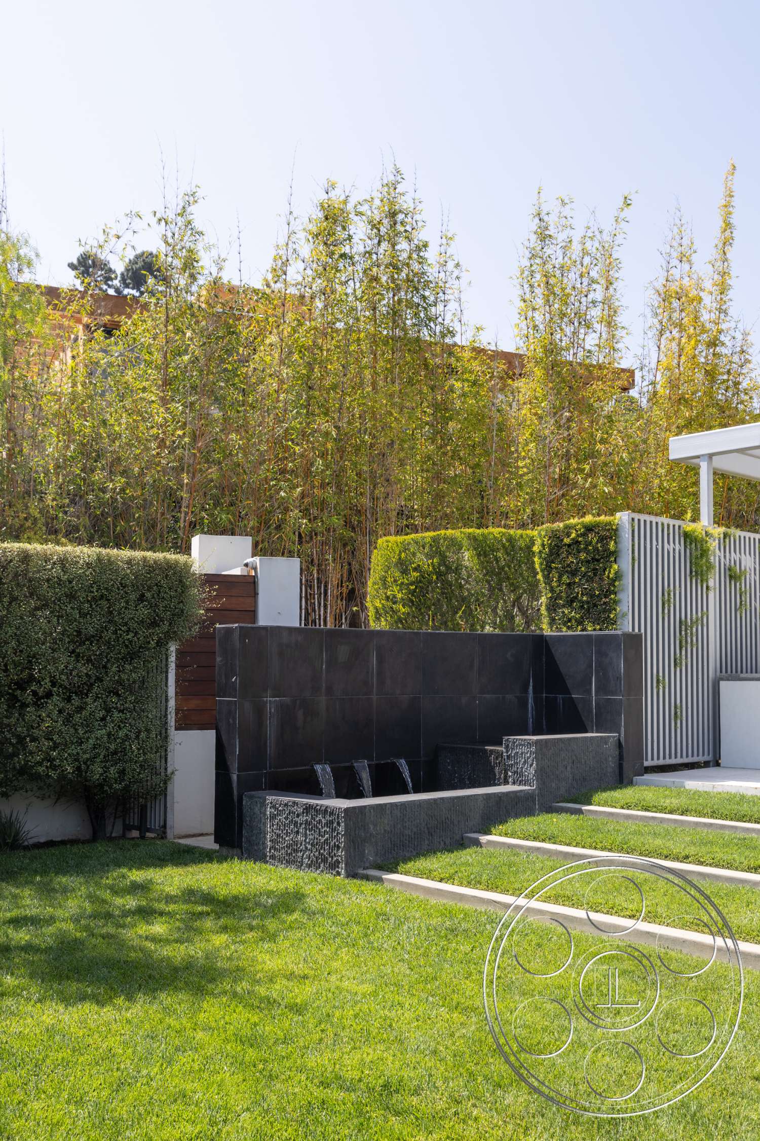 Modern 14 - exterior, wooden fence, symmetrical layout, concrete seating area, modern landscaping design, black stone water feature wall, steps with concrete borders, well-manicured grass lawn, tall bamboo privacy plants, horizontal metal slats