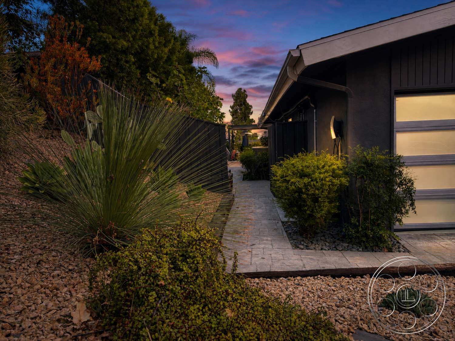 Americana 435 - exterior, lush greenery, concrete pathway, stucco exterior, sloping hillside, natural stone gravel, nighttime lighting, black wooden fence, landscaped garden beds, textured concrete pavers, flat roofline, modern house design, low-maintenance drought-tolerant plants