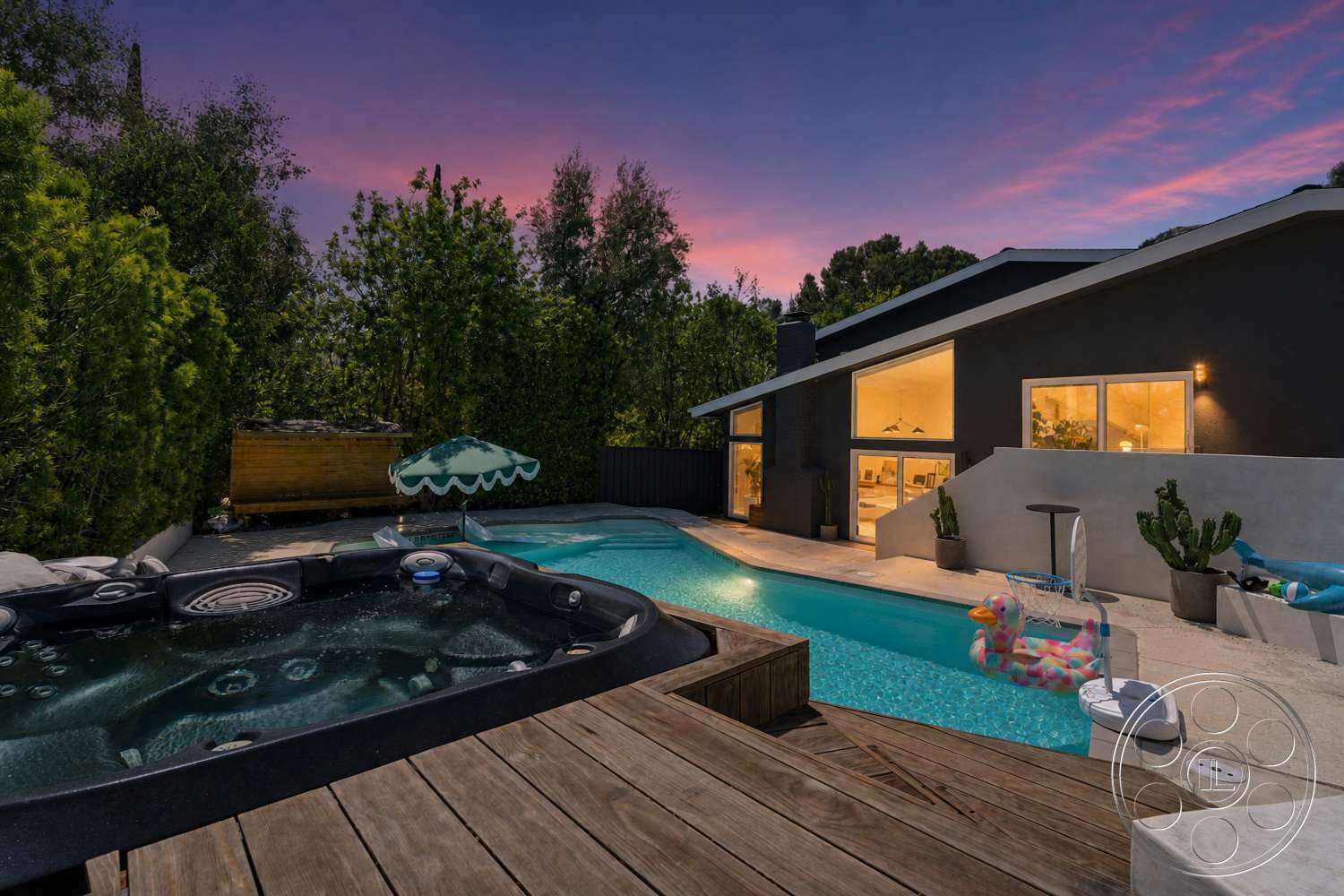Americana 435 - exterior, rectangular swimming pool, floor-to-ceiling windows, sloped roof, modern architectural style, patio seating area, lush green landscaping, outdoor umbrella, stucco exterior walls, potted cacti, concrete pool decking, wooden deck framing, fiberglass hot tub, poolside float toys, evening twilight sky