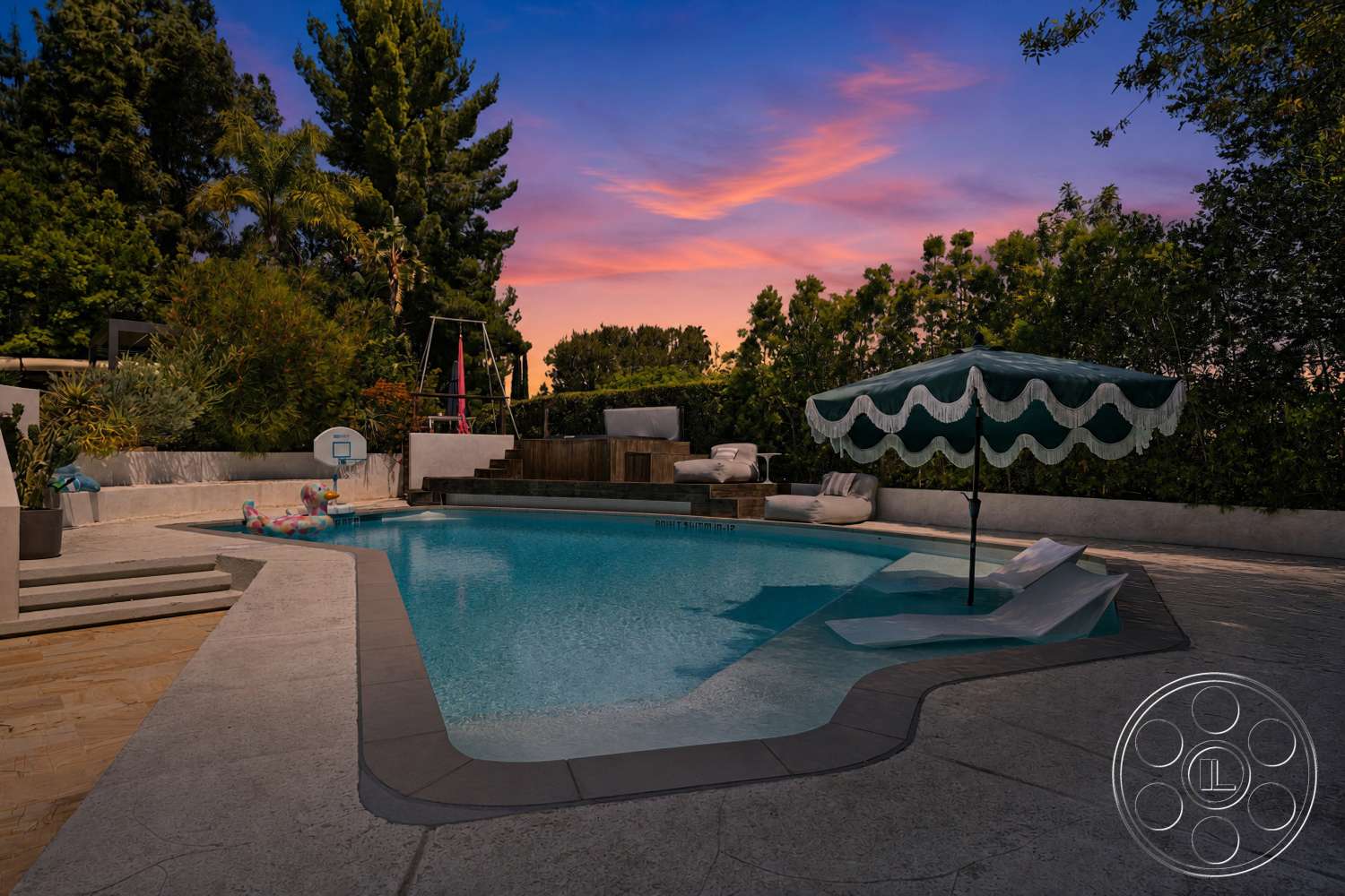 Americana 435 - exterior, outdoor seating area, rectangular swimming pool, concrete pool deck, landscaped garden area, wooden deck area, lush greenery surrounding pool, concrete patio steps, swimming pool with poolside lounge chairs, fiber umbrella with scalloped edge, evening sunset sky, kid-friendly pool features, playground swingset