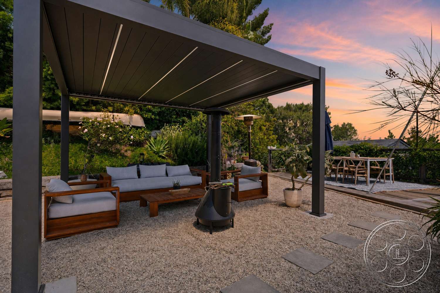 Americana 435 - exterior, wooden dining table, outdoor dining area, tropical landscaping, decorative potted plants, lush greenery backdrop, wooden outdoor furniture, stepping stone pathway, fabric upholstered seating, modern patio structure, metal pergola roof, natural gravel flooring, stone outdoor fire pit, aluminum patio heater, serene sunset view