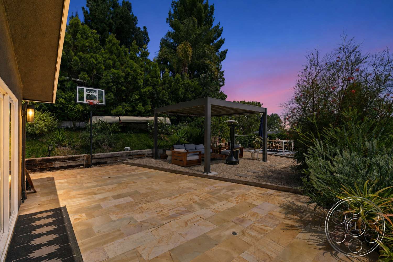 Americana 435 - exterior, green foliage, basketball hoop, stone retaining wall, outdoor heater, landscaped garden setting, metal pergola structure, sandstone patio flooring, wood outdoor seating, dusk sky view