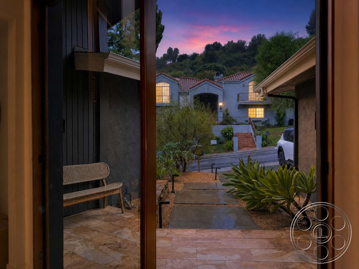 Americana 435 - large windows, exterior, suburban setting, mountain backdrop, sloped driveway, textured plaster walls, succulent plants, wooden front door, landscaped garden bed, multi-story home, paved concrete pathway, evening sky, stone entryway flooring, metal outdoor bench, red tile roof details