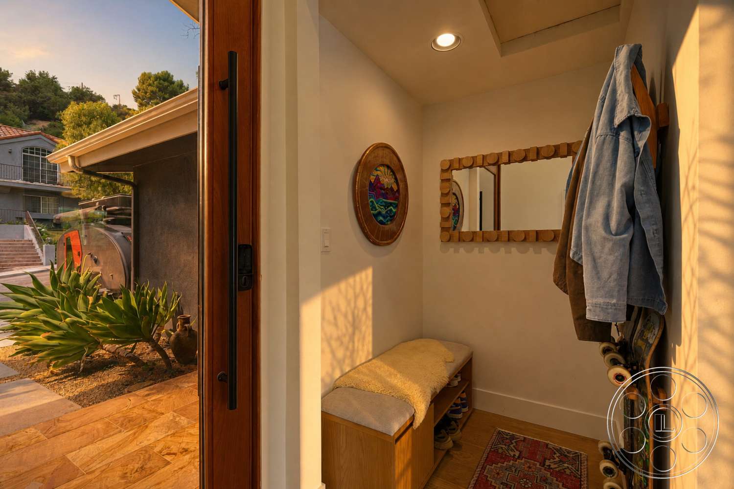 Americana 435 - hardwood flooring, decorative area rug, minimalist design elements, colorful wall art, textured plaster walls, wall-mounted coat rack, light-filled foyer, wooden entrance door, built-in wooden bench seating, natural light from entryway, framed mirror with circular wooden accents, suburban setting visible through entrance