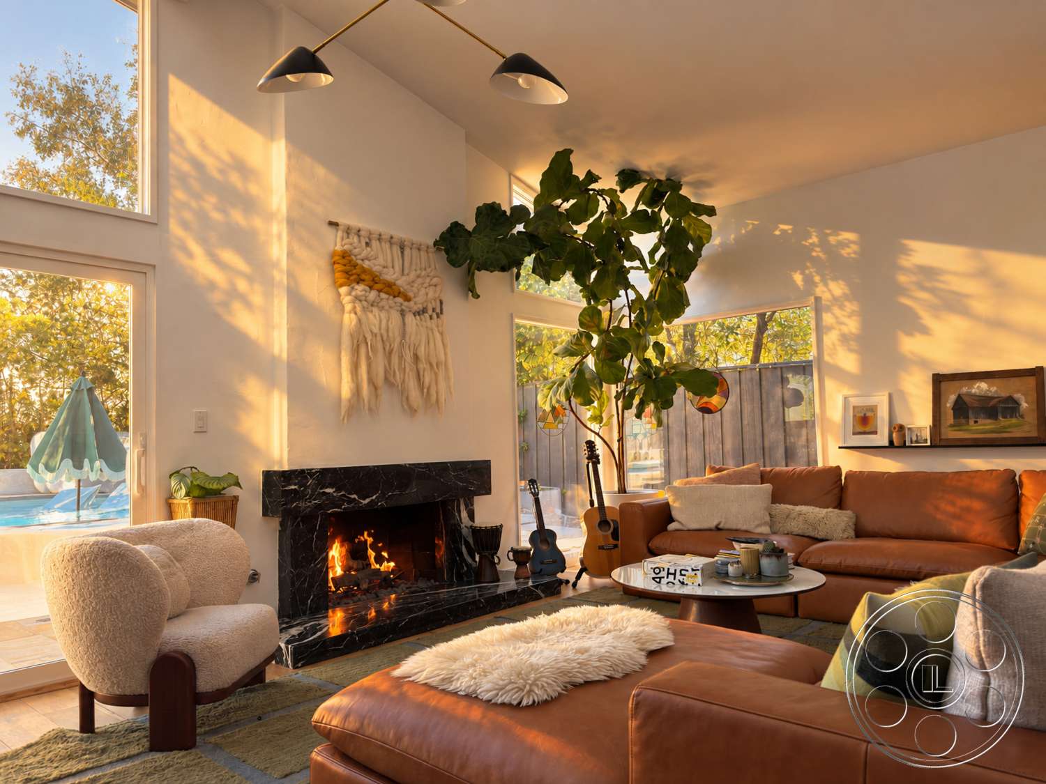 Americana 435 - natural light, wooden flooring, floor-to-ceiling windows, casual seating area, cozy living space, circular coffee table, white painted walls, large indoor plant, black marble fireplace surround, soft brown leather sectional sofa, textured wool wall hanging, framed artwork on shelves