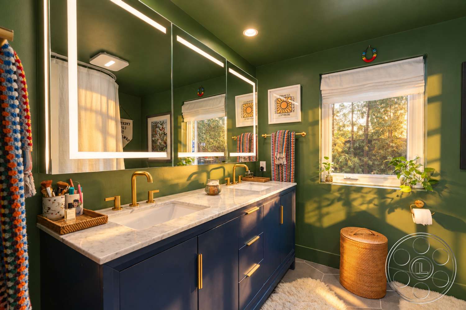 Americana 435 - potted plants, marble countertop, soft ambient lighting, framed wall art, woven storage basket, gold faucet fixtures, contemporary bathroom design, wall-mounted towel holder, rectangular undermount sinks, green painted walls, white mirror with LED backlighting, dark blue bathroom vanity, rectangular window with white roller shades, beige shag bathroom rug, circular woven stool