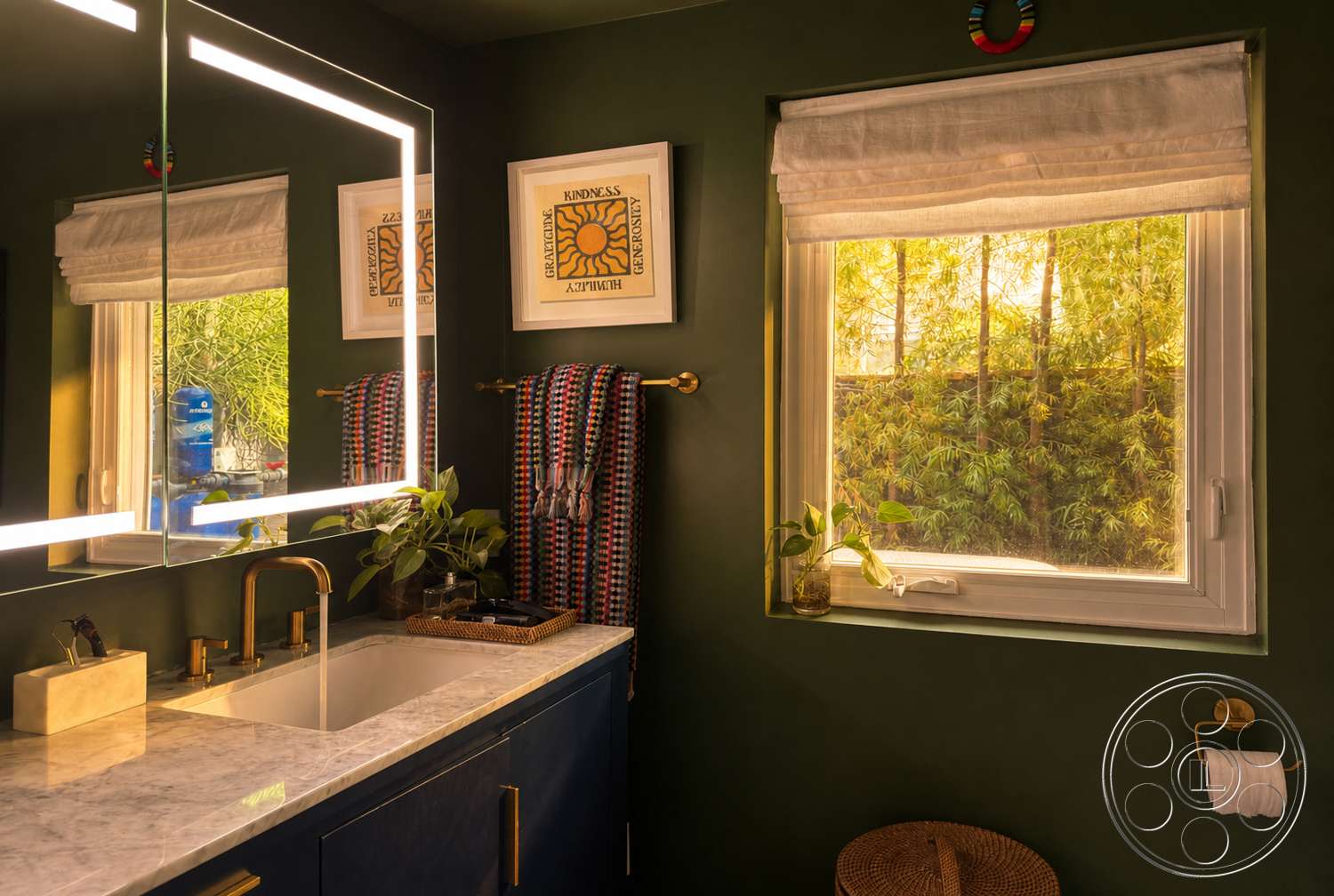 Americana 435 - towel rack, planted greenery, framed wall art, window with view of greenery, woven storage basket, modern bathroom design, marble bathroom countertop, green painted walls, dark blue vanity cabinet, gold faucet and fixtures, rectangular mirror with LED backlighting, roll-up window shades