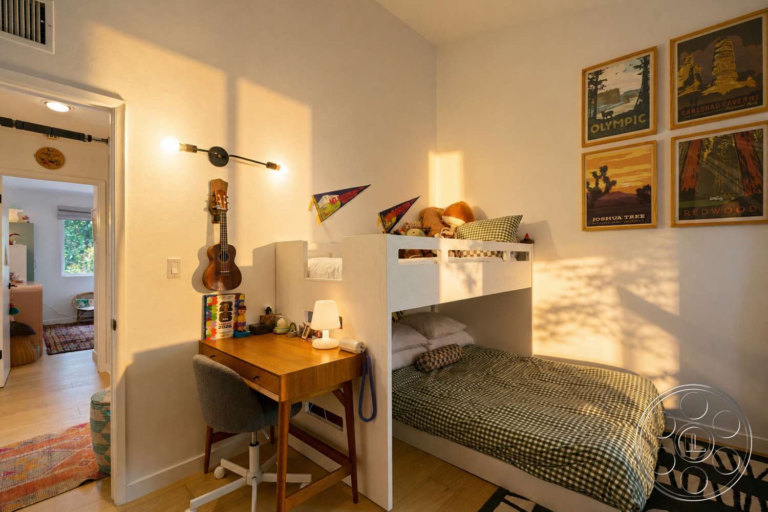 Americana 435 - light wood flooring, open doorway, black and white patterned area rug, natural light from window, white painted walls, wooden desk with drawers, colorful hanging decorations, loft-style bunk bed, gray upholstered desk chair, wall-mounted guitar hanger, framed nature-themed posters, table lamp with white shade, cotton checkered bedding