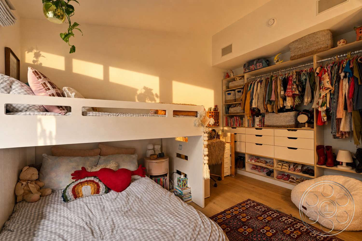 Americana 435 - textile accent pillows, child-friendly decor elements, hanging indoor plants, lofted bed with built-in storage, plush bedding with checkered patterns, soft wooden flooring, clothing storage shelves, curtained closet space, decorative area rug with floral pattern, wooden storage bins, multicolored stuffed toys, warm natural light from windows, cozy reading nook with books