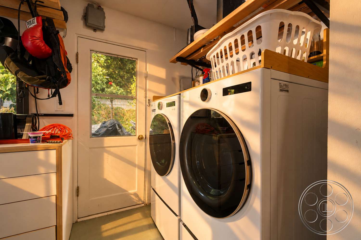 Americana 435 - laundry basket, white painted walls, light gray vinyl flooring, modern washer and dryer appliances, wooden countertop above appliances, glass door with metal handle, overhead wooden shelves, organizer hooks for bags, natural light from the door