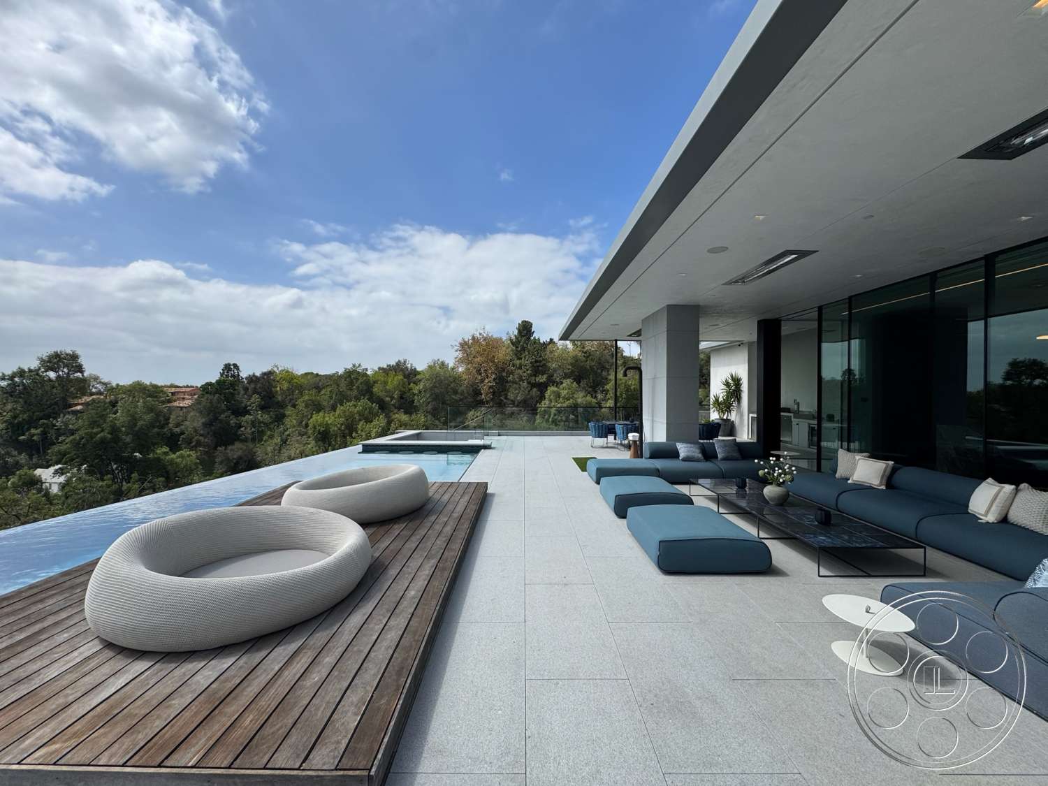 The Bellagio Estate - exterior, minimalist furniture, wood deck, city skyline view, infinity pool, rectangular pool, large sliding glass doors, modern outdoor lounge area, lush green landscaping, flat roof design, concrete patio flooring, oversized outdoor cushions