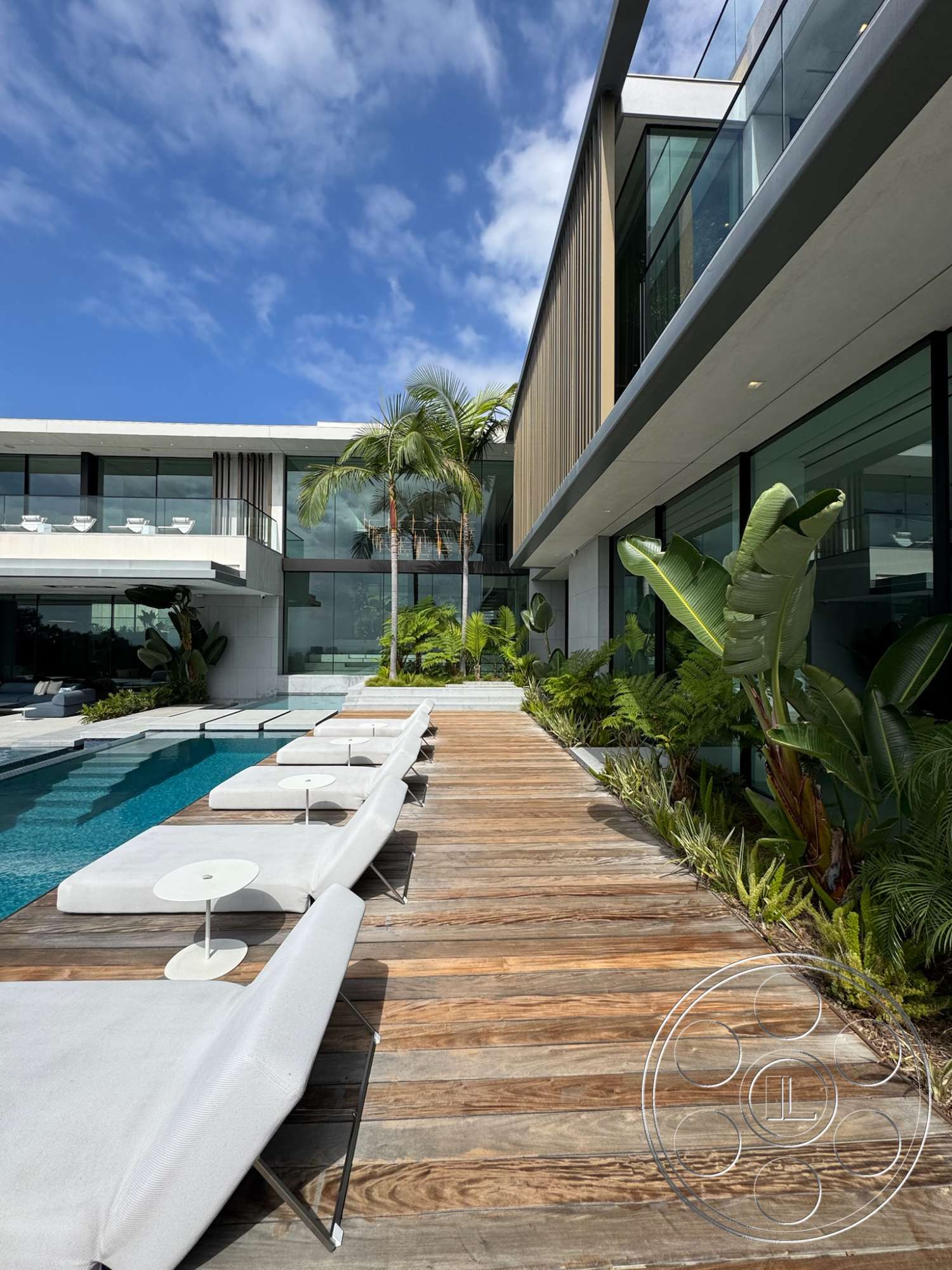 The Bellagio Estate - exterior, greenery accents, sliding glass doors, outdoor seating area, rectangular pool shape, modern lounge chairs, flat roof structure, palm trees landscaping, wooden deck pathway, glass wall facade, concrete swimming pool